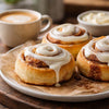 Ingredients for making cinnamon rolls laid out on a wooden surface with cinnamon sticks and brown sugar bowls.